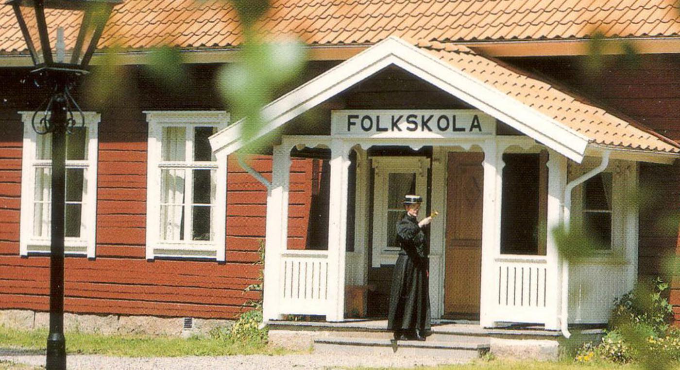 Tidsenligt klädd lärarinna på farstun till skolmuseets faluröda byggnad.