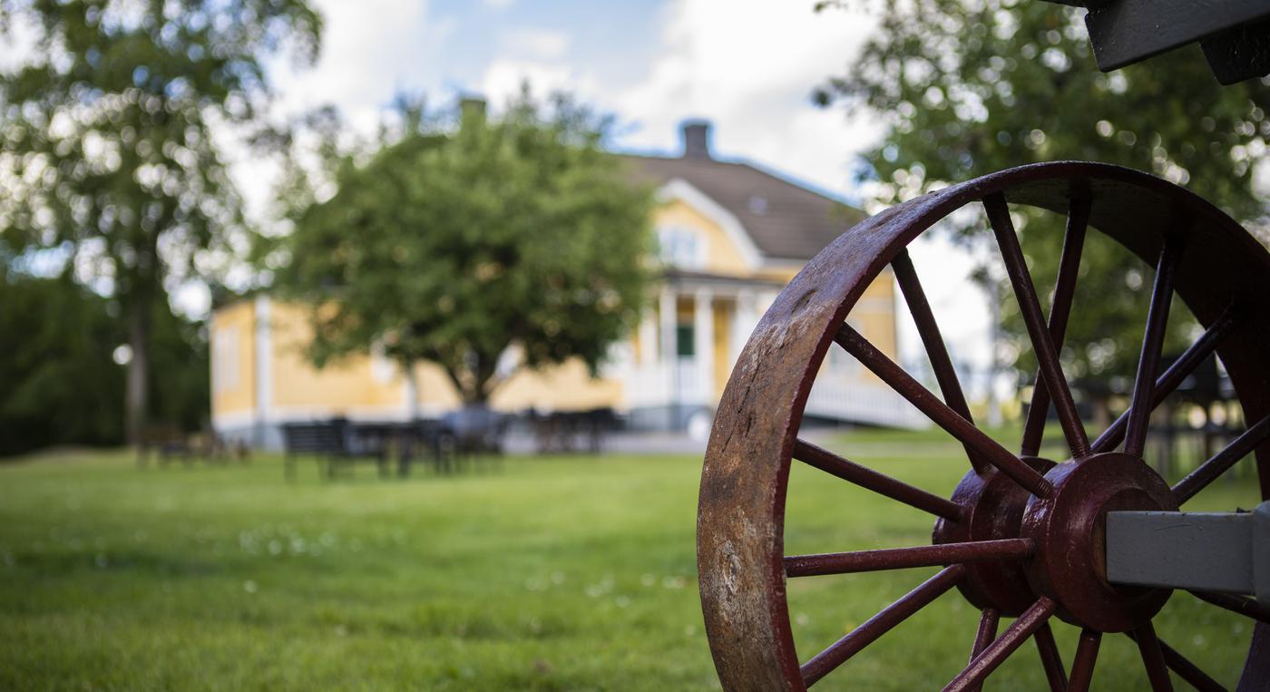 Ett rostigt hjul framför en gul byggnad.