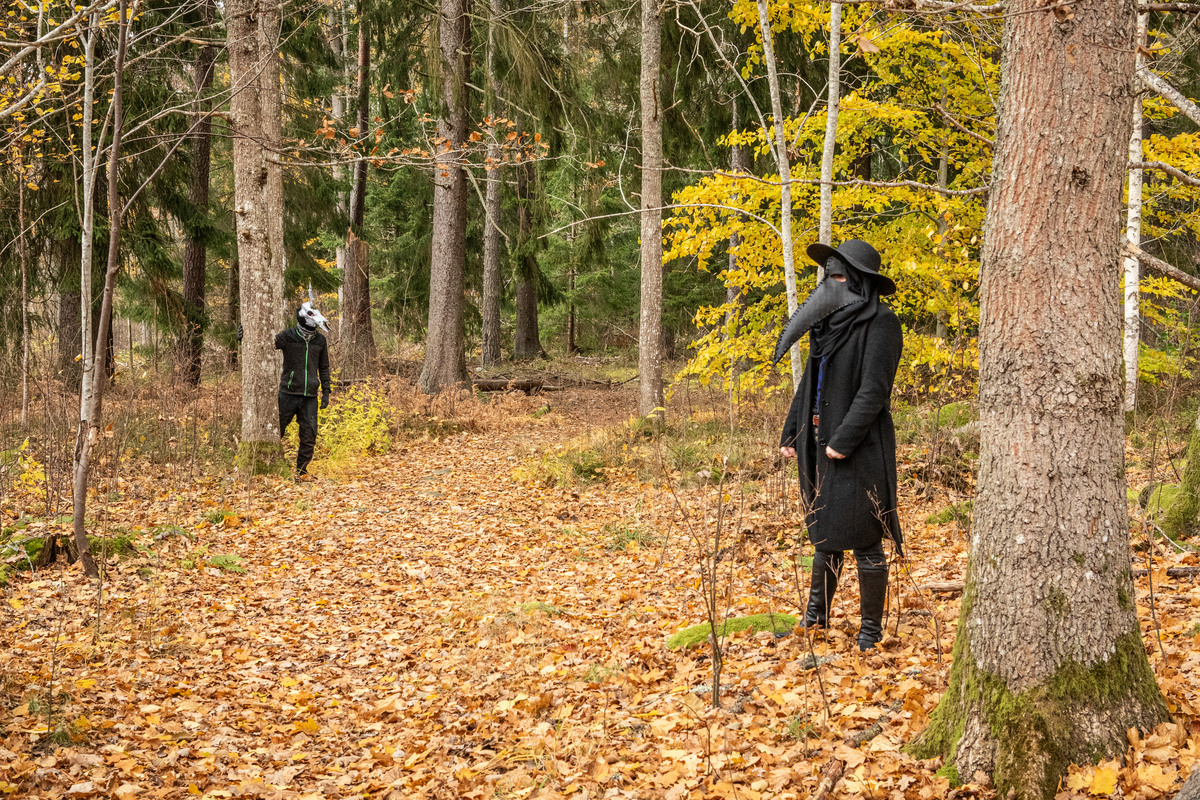 En person med ett djurkranium på huvudet och en med pestmask står längs en stig i skogen.