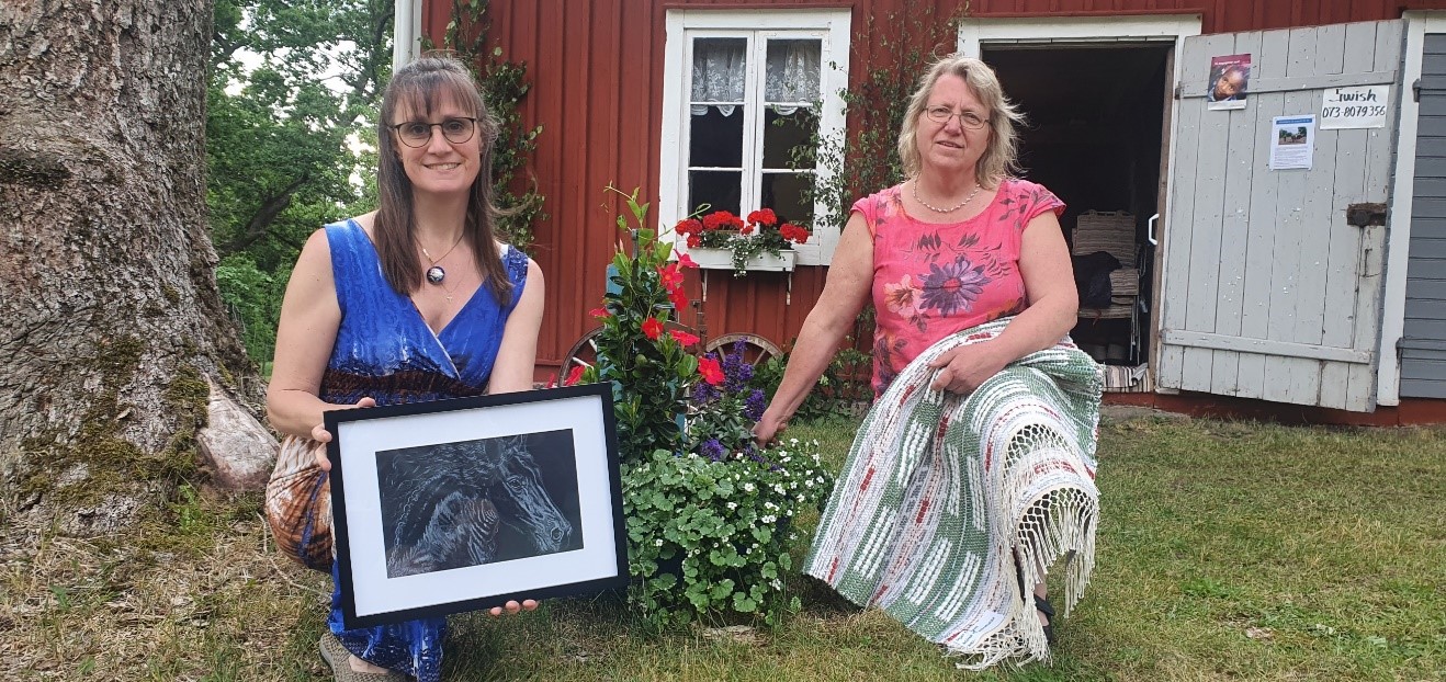 Katarina och Inger Eriksson sitter på huk framför det 100-åriga sädesmagasinet med varsitt egengjort alster i händerna.